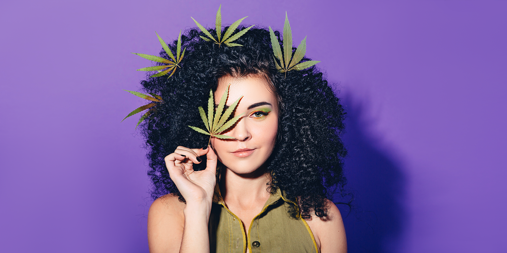 Woman with cannabis leaves in her Black Curly hair and holding up a cannabis leaf to her face